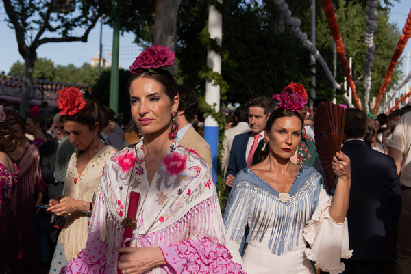 Women wearing traditional flamenco dresses, often in bright colors, and accessorized with flowers in their hair are seen laughing during the fair. The April Fair is one of Seville's most renowned and globally celebrated festivals. Originating in 1847 as a livestock fair, its festive spirit gradually overshadowed its commercial roots, transforming it into an indispensable event for Sevillians. For a week each year, the fairgrounds host over a thousand booths, serving as a second home for the city's residents. Attendees dress in traditional Andalusian attire, with men donning rural attire and women dressed in flamenco dresses.