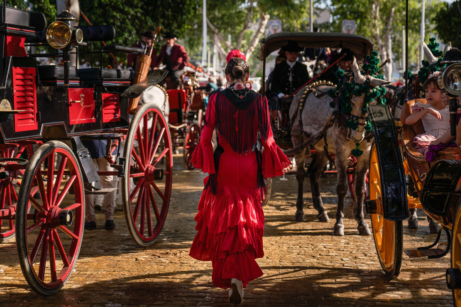 A woman wearing typical Sevillian costumes (round dresses) is seen walking in the fair. The April Fair is one of Seville's most renowned and globally celebrated festivals. Originating in 1847 as a livestock fair, its festive spirit gradually overshadowed its commercial roots, transforming it into an indispensable event for Sevillians. For a week each year, the fairgrounds host over a thousand booths, serving as a second home for the city's residents. Attendees dress in traditional Andalusian attire, with men donning rural attire and women dressed in flamenco dresses.