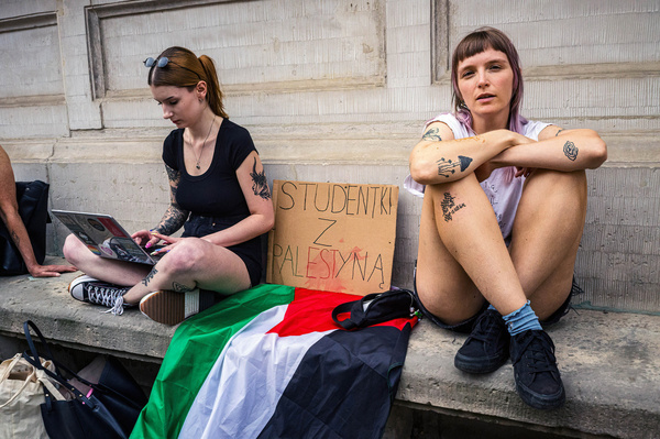 Two protesters sit together with a placard and a Palestinian flag between them. On Friday, May 25th, students at the University of Warsaw began a protest encampment on campus, opposing the university's ongoing relationships with Israeli academic institutions, research centers, and companies. They demand transparency regarding the university's partnerships and insist on cutting ties with entities linked to the occupation of Palestine and the ongoing conflict. On Tuesday, May 29th, representatives from the student encampment met with the Rector of the University to begin negotiations, while a smaller protest continued outside.
