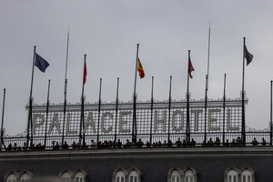 Several people watch the parade under a sign for the Palace Hotel. During the Armed Forces parade on National Day led by King Felipe VI, Queen Letizia, their daughters Leonor de Borbón, Princess of Asturias and Infanta Sofía de Borbón.
