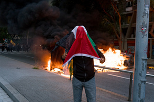 A pro-Palestinian protester is seen draped with a Palestinian flag as dumpsters burn in the background during the demonstration. A nationwide strike was held all over Spain, with demonstration in all the main cities. More than 20.000 of pro-Palestinian protesters marched in Barcelona, with several clashes with police and episodes of looting and burning of dumpsters.