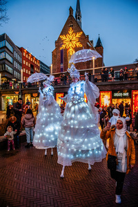 Two women on stilts are walking on the streets. The Winter Wonder Parade marks the festive opening of the annual Nijmegen Winter Weeks. This enchanting spectacle features a fairytale procession including a winter carriage pulled by white horses, ice queens on stilts, musicians, dancers, and jugglers. The parade departs from the Lindenberg at 4:30 PM and concludes at the Grote Markt, where the Christmas tree lighting officially starts three weeks of winter magic.