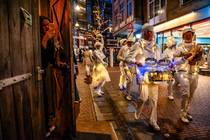 A woman is sneaking out of a pub to take a photo of the music band. The Winter Wonder Parade marks the festive opening of the annual Nijmegen Winter Weeks. This enchanting spectacle features a fairytale procession including a winter carriage pulled by white horses, ice queens on stilts, musicians, dancers, and jugglers. The parade departs from the Lindenberg at 4:30 PM and concludes at the Grote Markt, where the Christmas tree lighting officially starts three weeks of winter magic.