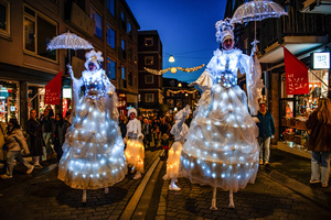 Two female acrobats on stilts are performing in the middle of the street. The Winter Wonder Parade marks the festive opening of the annual Nijmegen Winter Weeks. This enchanting spectacle features a fairytale procession including a winter carriage pulled by white horses, ice queens on stilts, musicians, dancers, and jugglers. The parade departs from the Lindenberg at 4:30 PM and concludes at the Grote Markt, where the Christmas tree lighting officially starts three weeks of winter magic.