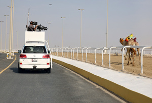 A live television camera captures the start of the purebred Arabian camel race at the Al Shahaniya Camel Racing Track. Camels are guided by operators using wireless devices that control robot jockeys, pulling the reins and encouraging the animals through built-in loudspeakers. In 2005, Qatar banned child jockeys from camel racing following international concerns over child exploitation and trafficking. To preserve the sport, lightweight robot jockeys were introduced and are remotely operated by trainers driving alongside the track.