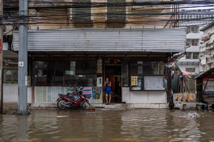 A person stands in the doorway of a building as floodwater covers the street outside near Phetchaburi Road. Floodwaters inundate major roads, including Phetchaburi Road and the Pridi Banomyong neighborhood in Bangkok, following a record monsoon downpour that prompted a capital-wide disaster declaration. Municipal pumps operate at full capacity to drain overflowing canals.
