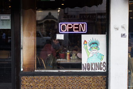 A neon “OPEN” sign is displayed in a restaurant window with diners seated inside, and a “NO KINGS” poster visible in the window during the No Kings protest.