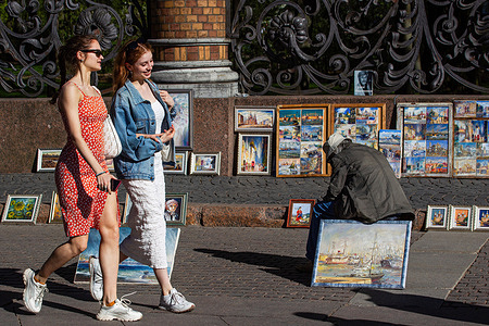 Young women walk along the street of the center of St. Petersburg as an artist sits in front of his paintings on a summer day.