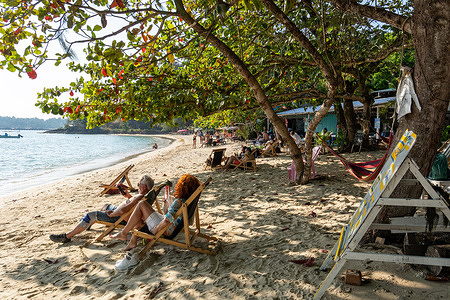 A view of a tourist couple sitting on beach chairs under a tree at Klong Kloi Beach on Koh Chang Island, Thailand. Koh Chang Island is Thailand’s third-largest island after Phuket and Samui. It is located around 300 kilometres east of Bangkok in Trat Province, in the Gulf of Thailand, near the Cambodian border. January falls within the peak tourism season, as it is part of the cool, dry period when weather conditions are most favourable, and visitor numbers are at their highest.