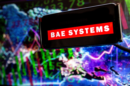 In this photo illustration, the BAE Systems logo is seen displayed on a mobile phone with a world map and stock graph illustration on a computer screen in the back ground.