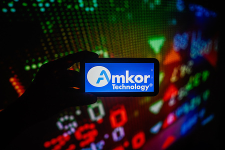 In this photo illustration, an Amkor Technology logo is displayed on a smartphone with stock market percentages in the background.