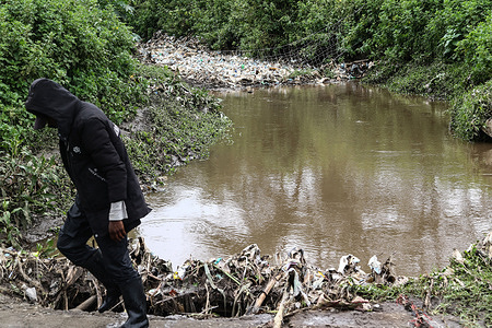 A man wearing a hooded jacket walks past a river polluted with plastic drink bottles in Nakuru. Plastic waste remains a big environmental nightmare in Kenya. In an attempt to address the problem, the government is planning to introduce an extended producer responsibility law to compel companies to be responsible for the end of life of their products.