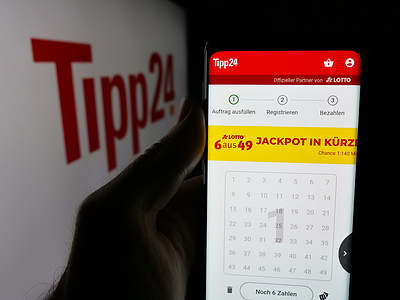In this photo illustration, a person is holding a smartphone with the webpage of German online lottery company Tipp24.de on screen in front of logo.