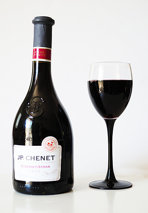 A bottle of J.P. Chenet Cabernet-Syrah French red wine next to a filled wine glass with a black stem on a white surface.