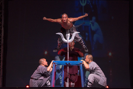 A shaolin participant seen laying on a big spear at a show.
Shaolin The Legend is a group of Shaolin Kung Fu Masters. They fly in the air, impress with their swords, and break stones in their body or head and their bodies touch on spears, all in a dull atmosphere with lighted candles and stunning scenery.