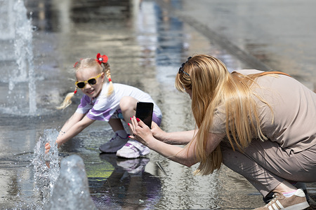 A woman seen recording a young girl as she plays in a city fountain to cool off during a hot summer day. A hot summer day forced the city to seek refuge from the heat in the coolness of its fountains. In such moments, the squares fill with children's laughter as kids joyfully play among the refreshing jets of water trying to find the desired coolness amidst the scorching asphalt. This is a time of relaxation.