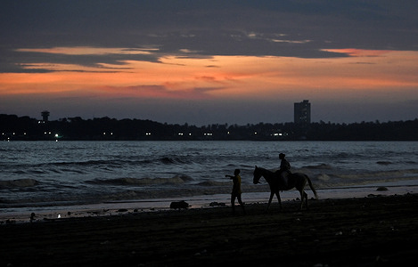 A woman enjoys a horse ride on a beach during the sunset in Mumbai. High humidity post monsoon season and tiny water droplets acts like a prism, reflecting and refracting sunlight which in turn enhances red and orange hues making the sky look beautiful as the sun sets in the evening.