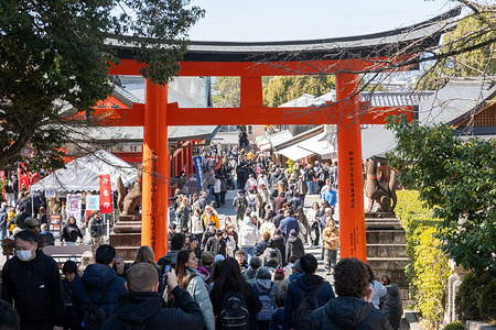 Visitors walk through a large vermilion torii gate along the busy approach to Fushimi Inari Taisha in Kyoto, Japan, with shops and crowds lining the pathway near the shrine entrance. Fushimi Inari Taisha is a famous Shinto shrine in Kyoto known for its thousands of bright red torii gates that form winding paths up the mountainside. Dedicated to the god of rice and prosperity, it’s one of Japan’s most iconic and heavily visited religious sites.