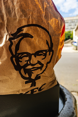 A crumpled brown bag with a black KFC Colonel Sanders logo on it, sticking out of the trash can, with a blurred cityscape in the background.