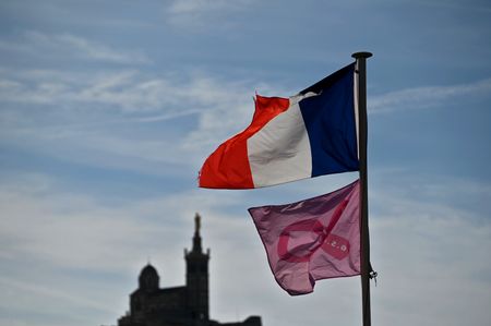 A pink flag adorned with a ribbon, a signature symbol associated with "Pink October", flies beneath a French tricolor flag (blue, white, and red), with the silhouette of the Notre-Dame de la Garde basilica in Marseille visible in the background. For "Pink October", a month dedicated to raising awareness about breast cancer screening, the city of Marseille is decked out in pink, with flags on display, as seen here at the Old Port (Vieux-Port).