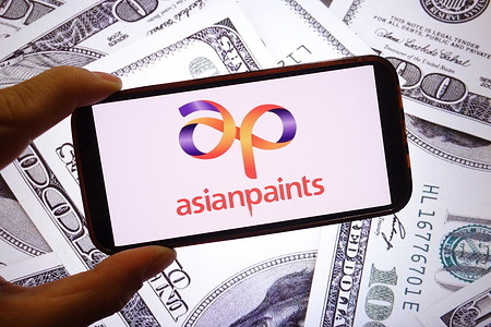 In this photo illustration, the Asian Paints company logo is seen displayed on a smartphone screen with United States dollar banknotes in the background.