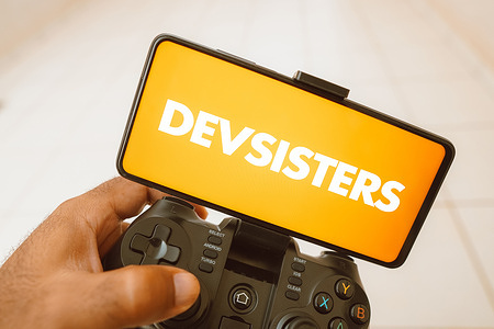 In this photo illustration, a person plays on a joystick and the Devsisters Corporation logo displayed on a smartphone screen.