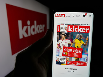 In this photo illustration, a person is holding a cellphone with the webpage of German football and sports magazine Kicker on screen in front of logo.