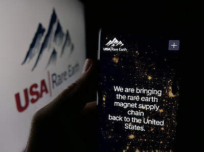 In this photo illustration, a person is holding a cellphone with the webpage of US mining company USA Rare Earth Inc. on screen in front of logo.