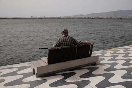 A man is seen fishing while sitting on a bench near the sea.