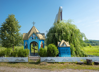 A quaint chapel with vibrant wall art is seen in Teresva, accompanied by a nearby stork sculpture and a refreshing well that adds charm to the serene landscape on a sunny day. Chapel with Stork and Well (Teresva Village, Transcarpathia).
This unique cult and ethnographic complex is located near the village of Teresva in Transcarpathia. It includes a brightly decorated blue-and-gold chapel, above which stands a large sculpture of a white stork. The stork is one of the most important symbols of Ukrainian culture, embodying peace, prosperity, and family comfort. The presence of the stork next to the chapel reflects a deep syncretism of local Christian and folk beliefs. On the complex's grounds, under a willow tree, there is a well, emphasizing its practical and symbolic significance for the local community. The site is a harmonious blend of spiritual traditions and folk life within the rural landscape of Transcarpathia.