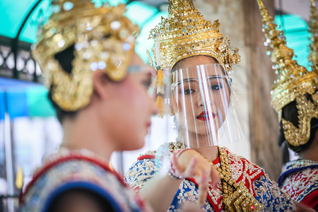 BANGKOK, THAILAND - MAY 04, 2020: Thai female dancers wearing traditional dress and face shields to protect themselves against Coronavirus (Covid-19) dancing at the Erawan Shrine in Ratchaprasong Junction.