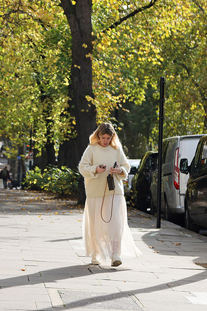 A woman strolls past trees glowing with autumn colours under the sunshine, in London.
