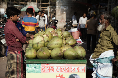 Two men talk business over a coconut seller's stall. 
Dhaka, the capital of Bangladesh lies along the east bank of the Buriganga River in the heart of the Bengal delta. The city is a microcosm of the entire country, with diverse religious and ethnic communities. Dhaka is the economic, cultural and political center of Bangladesh and is a major financial center of South Asia. It is one of the world's most populated cities with a population of 18.89 million people in the Greater Dhaka Area.