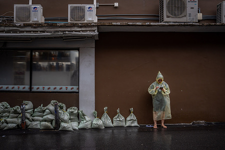 A woman wearing a rain poncho checks her phone beside sandbags placed outside a building in the Ekkamai area of Bangkok. Floodwaters inundate major roads, including Phetchaburi Road and the Pridi Banomyong neighborhood in Bangkok, following a record monsoon downpour that prompted a capital-wide disaster declaration. Municipal pumps operate at full capacity to drain overflowing canals.