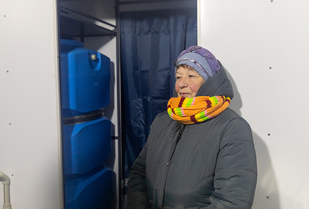An elderly resident seen inside a mobile Solidarity Point amidst the energy crisis. Large blue water tanks are seen stored on shelves behind the woman. A specialized mobile assistance point, the "Solidarity Point" has begun operating to support residents who have been without electricity, heating, and water for weeks following massive strikes on infrastructure. The mobile complex consists of several units one where citizens can warm up, obtain hot water, charge their devices, and another equipped with washing machines and dryers to address basic hygiene needs. This initiative aims to restore essential living conditions during the severe energy crisis by adapting technical solutions previously developed for frontline support to meet civilian needs. Given the high demand for these services, the project is set to scale up and deploy mobile stations to other districts of the capital where the utility situation remains most critical.