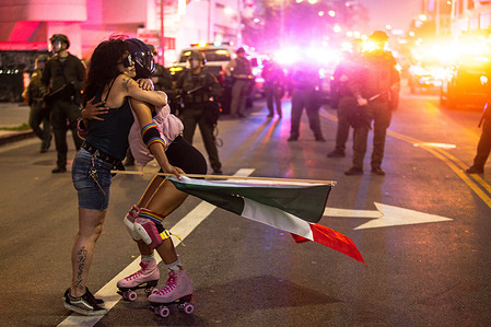Two demonstrators embrace in the middle of the street as riot police and bright lights fill the background, during an anti-ICE protest. A demonstration staged as part of a national day of action drew hundreds of protesters who marched against ICE and police violence in downtown Los Angeles