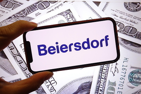 In this photo illustration, the Beiersdorf company logo is seen displayed on a smartphone screen with United States dollar banknotes in the background.