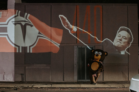 A projected image is cast onto the side of a building along Santa Monica Boulevard as a costumed person walks beneath the projection during a Tesla Takedown demonstration outside the Tesla Diner.