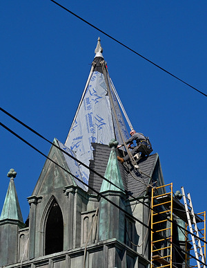 A steeplejack renovates the shingles on a church steeple under blue skies.
