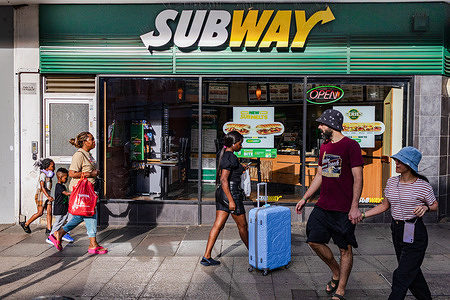 People walk past a Subway restaurant in London. Subway IP LLC, is an American multinational fast-food restaurant franchise that specializes in submarine sandwiches, wraps, salads, and drinks.