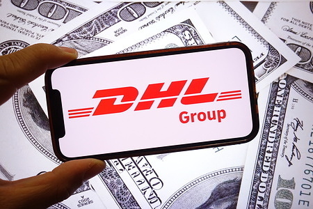 In this photo illustration, the DHL Group company logo is seen displayed on a smartphone screen with the United States Dollar banknotes in the background.