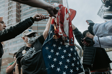 Protesters hold and ignite a United States flag using handheld lighters during an anti-ICE demonstration near the Metropolitan Detention Center in downtown Los Angeles. Several individuals take turns applying flame to the fabric while others document the moment with phones and cameras. The protest targeted U.S. Immigration and Customs Enforcement and federal immigration detention practices, drawing activists to the area surrounding the detention center.