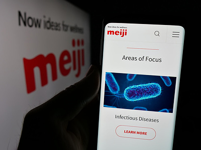In this photo illustration, a person is holding a cellphone with the webpage of Japanese business Meiji Holdings Company Ltd. on screen in front of logo.
