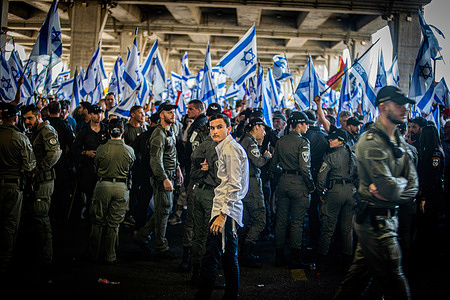 An Ultra-orthodox Jewish youth looks back as he stands behind a line of border police officers facing protestors waving the Israeli flag during a demonstration at the Ben Gurion international airport. Israeli police have clashed with protesters amid demonstrations against the government's controversial judicial reform plans. Protests broke out after a bill to remove the power of the Supreme Court to review ministers' decisions passed its first reading in parliament on Monday night. The judicial reform has polarized the country, sparking months of mass demonstrations.