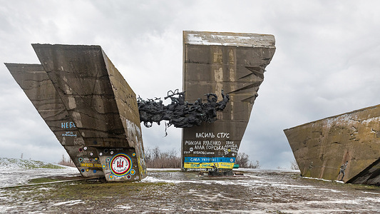 The damaged central structure of the Memorial of Glory in Izyum is seen featuring three massive concrete slabs. Impact marks and cracks are seen on the gray surfaces. Pro-Ukrainian graffiti, including a blue and yellow flag and names, is seen painted over the war-torn monument. The Memorial of Glory on Mount Kremianets remains one of the most significant testaments to the destruction of cultural heritage in the Kharkiv region. The central part of the complex—the "Attack" monument—remains in a semi-ruined state: one of the concrete stelae, toppled in 2022, lies at the base of the monument. International investigative teams have officially recorded the damage to the site as a war crime. Although full restoration has been postponed until the end of active hostilities, the memorial remains open to visitors and continues to serve as a place of remembrance for the victims of two wars.