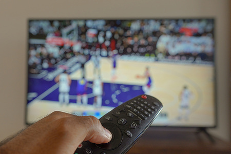 In this photo illustration, a hand holding a TV remote control seen in front of a TV broadcasting a basketball match.