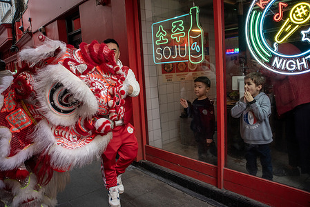 Two children look at the lion through the window before it enters the shop in Chinatown, London. For Chinese New Year (Lunar New Year) a lion dance group performed at shops and restaurants to bring good luck, fortune, and prosperity while driving away evil spirits. The performance involves the lion "eating" greens (lettuce/cabbage) and red envelopes (hongbao) offered by business owners.