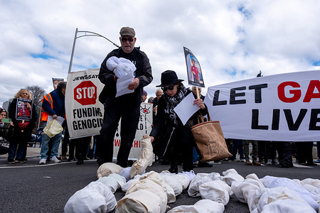 Two protesters lay down effigies of dead Palestinian children wrapped in white cloth. A small group of protesters gathered at the end of the street where Senator Chuck Schumer lives to demand an immediate end to all US funding to Israel. Led by activist group Jewish Voice for Peace, the group walks through a farmer's market along Prospect Park carrying banners and effigies of Palestinian children killed in Gaza by Israeli bombing in the Israeli-Hamas War.