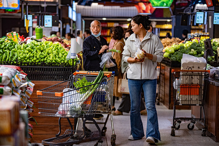 A consumer seen shopping at Hualian Supermarket. She is holding an item and there is a shopping cart with vegetables beside her.