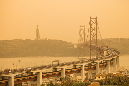 View of the Ponte 25 de Abril Bridge and Cristo Rei statue amidst the orange sky in Lisbon. The phenomenon, known as "Clay rain", is related to the transport of sand particles from the Sahara desert to the Iberian Peninsula and affects the interior North and Centre of Portugal.